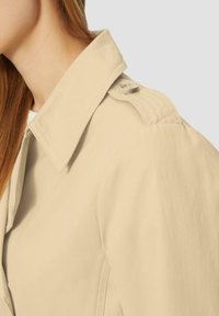Beige trenchcoat met een brede kraag, knoopsluiting op de epauletten en een gladde textuur, met zichtbare stiksels langs de mouwen.
