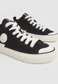 Černé plátěné tenisky s bílými plochými tkaničkami, gumovou špičkou a texturovanou bílou podrážkou. Na boku se nachází kruhové logo.