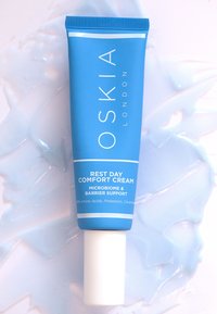Blaues Tubenpackung der Oskia Rest Day Comfort Cream, mit weißem Text, glatter Textur und abgerundetem Deckel. Der Hintergrund zeigt eine hellblaue Cremetextur.