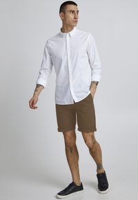 Chemise blanche à manches retroussées, associée à un short marron. Des baskets noires complètent la tenue. Tissu lisse, design décontracté.
