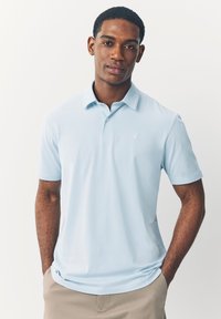 Šviesiai mėlyna polo marškinėlių su minkšta medžiaga, su apykakle, trumpomis rankovėmis ir mažu išsiuvinėtu logotipu ant krūtinės.