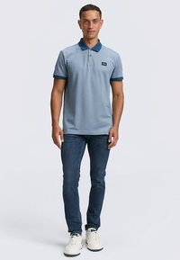 Világoskék galléros póló sötétkék gallérral és mandzsettával, sötétkék farmerrel és fehér tornacipővel viselve. Bal oldali mellrészen címkével.