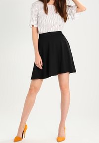 Schwarzer A-Linien-Rock mit schmaler Taille, kombiniert mit einer weißen Bluse mit Polka-Dots. Verfügt über kurze, geraffte Ärmel. Trägt dazu gelbe High Heels.
