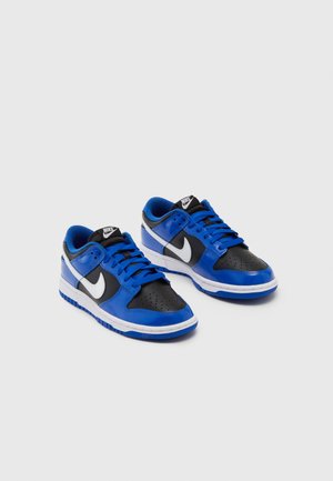 Paire de baskets Nike basses en bleu, noir et blanc avec des logos swoosh blancs, des lacets bleus et des semelles intermédiaires blanches sur fond gris.