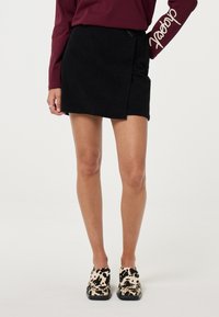 Zwarte corduroy mini-jurk met een wikkeldesign, textuurstof en zichtbare zijkoord. Gecombineerd met slip-on schoenen met een dierenprint.