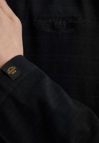 Camisa de manga larga negra con bordado de logotipo texturizado y etiqueta de marca en el puño, que muestra una confección de tela acanalada.