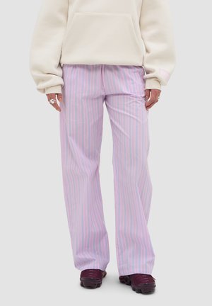 Person, die lockere pink gestreifte Hose, beige Kapuzenjacke, silberne Ringe und dunkelviolette Sportschuhe trägt, steht vor einfarbigem Hintergrund.