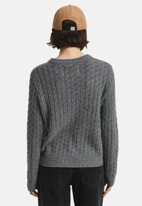 Grauer Kabelstrickpullover mit Rundhalsausschnitt und geripptem Saum, von hinten präsentiert, kombiniert mit dunklen Jeans und einer braunen Kappe.
