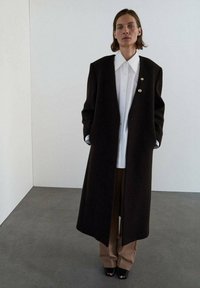 Abrigo largo de color marrón oscuro con solapas anchas y dos botones dorados, combinado con una camisa blanca y pantalones beige de pierna ancha. Textura suave, diseño estructurado.