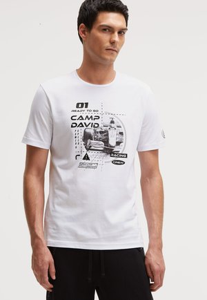 Junger Mann trägt ein weißes T-Shirt mit einem schwarzen Rennwagen-Grafik und dem Text "CAMP DAVID" und "RACING CREW" auf der Vorderseite.