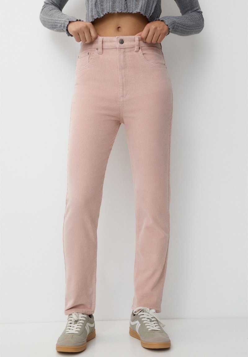 Personne portant un pantalon en velours côtelé rose clair taille haute et des baskets grises, avec un pull court gris côtelé, debout contre un mur blanc.