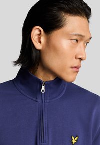 Marinblå zip hoodie med hög krage, strukturerat tyg och en gul broderad logotyp på vänster bröst. Täta profilbild.