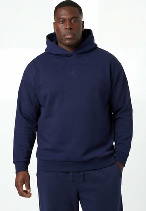 Dorosły mężczyzna w granatowej bluzie z kapturem i dopasowanych spodniach dresowych stojący na tle jednolitego białego tła.