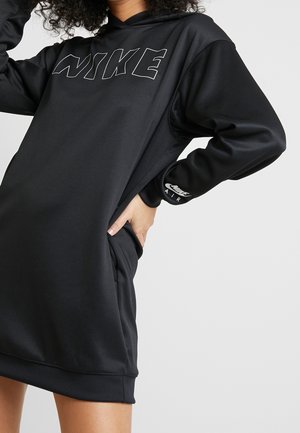 Osoba nosící černé šaty s potiskem Nike, s nápisem NIKE na hrudi a logem Nike Air na manžetě rukávu, s rukou v kapse.