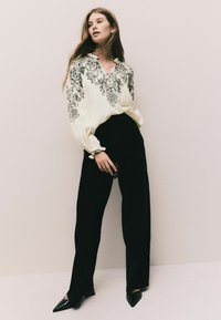 Bluză albă cu broderie florală neagră, mâneci lungi tip balon, asortată cu pantaloni largi negri cu talie înaltă și pantofi cu toc subțire negri.