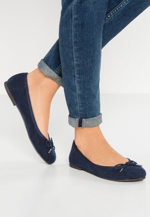 Navy blue ballet flats with a decorative bow worn with rolled-up blue denim jeans on light background.