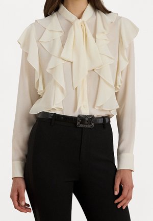 Woman wearing cream ruffled blouse with front tie and black pants with a decorative square-buckled black belt, standing with hands relaxed.