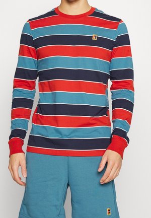 Person iført en langærmet skjorte med horisontale røde, blå og marineblå striber og turkis shorts med små logo-patches.