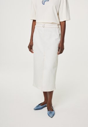 Vrouw draagt een crèmekleurige midi-rok met zichtbare naden en hartvormige riemdetails, gecombineerd met lichtblauwe slip-on schoenen met puntige neus.