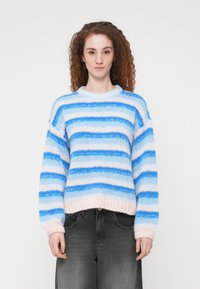 Γυναίκα με σγουρά κόκκινα μαλλιά που φοράει μπλε και λευκό ριγέ πλεκτό πουλόβερ και σκούρο γκρι τζιν μπροστά σε λευκό, απλό φόντο.