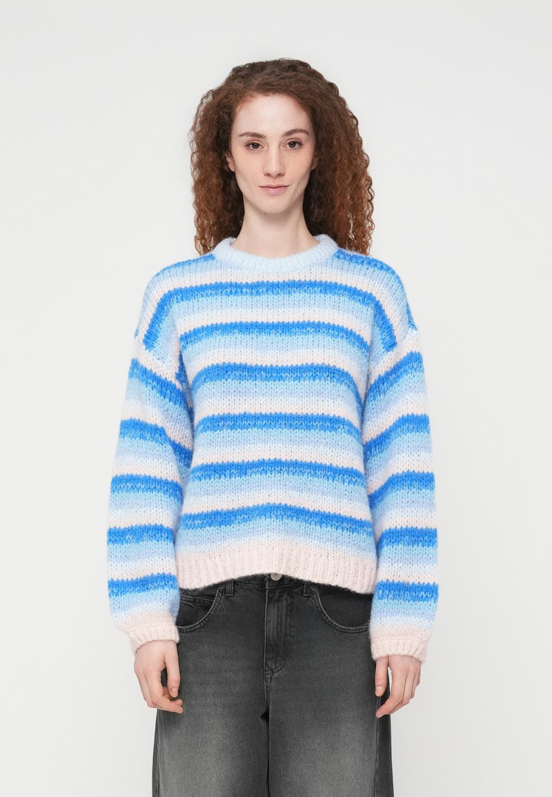 Γυναίκα με σγουρά κόκκινα μαλλιά που φοράει μπλε και λευκό ριγέ πλεκτό πουλόβερ και σκούρο γκρι τζιν μπροστά σε λευκό, απλό φόντο.