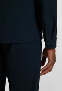 Camicia blu navy con tessuto testurizzato e maniche lunghe, dotata di bottoni ai polsini e vestibilità rilassata. Una mano poggia vicino alla cucitura della camicia.