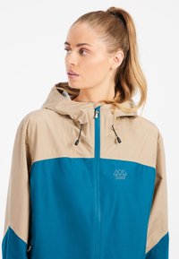 Tyrkysovo-hnedý vetrový kabát so zipsom, nastaviteľnou kapucňou a logom na hrudi. Hladký povrch a ľahký materiál.