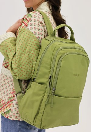 Kvinde med en lysegrøn nylonrygsæk med flere lynlåslommer over en bluse med blomsterprint i rødt og grønt patchwork.