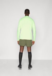 Neon žalia ilgomis rankovėmis palaidinė, alyvuogių žalios šortai, juodos kelio aukščio kojinės ir juodi sportiniai batai; glotni tekstūra ir sportiškas siluetas.