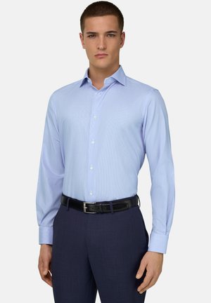 Mann trägt ein hellblaues, langärmeliges Hemd mit Knöpfen, das in dunkle marineblaue Hose mit schwarzem Gürtel gesteckt ist.