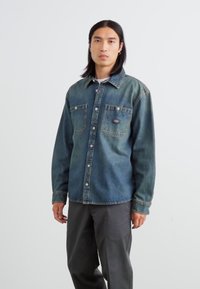 Mørkeblå denimskjorte med knapper, to frontlommer og kontrasterende syninger. Bruges over en hvid skjorte og er parret med grå bukser.