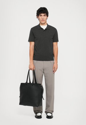 Bolso tote de cuero negro con doble asa, posicionado frente a un modelo que lleva una camiseta tipo polo gris oscuro y pantalones de pliegues gris claro.