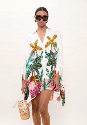 Mujer con vestido camisa blanco oversize con estampado colorido de palmeras tropicales, gafas de sol grandes, pendientes de aro dorados y llevando un bolso tejido.