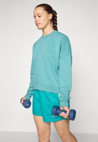 Teal sweatshirt med långa ärmar och en lila logotyp, tillsammans med mönstrade teal-shorts. Håller blå hantlar, står på en ljus bakgrund.