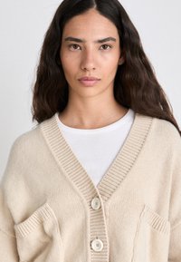 Beige knit cardigan with a V-neck, two front pockets, and two large buttons, layered over a white textured top. Soft, relaxed fit.