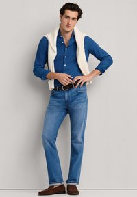 Muž v modrej košeli na gombíky, svetlomodrých džínsoch, hnedom opasku a mokasínach, s bielym svetrom prehodeným cez plecia, stojaci pred jednoliatym pozadím.