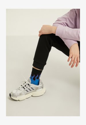 Chaussure de sport argentée avec une tige en mesh et une semelle en caoutchouc, associée à des chaussettes noires ornées de motifs de flammes bleues et violettes, portées avec un jogging noir.