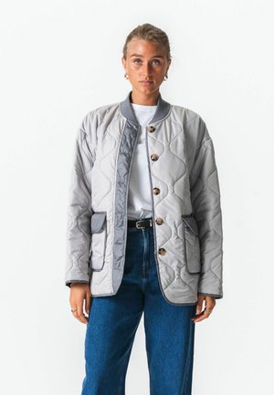 Jonge vrouw die een lichtgrijs doorgestikt jack met houten knopen, een wit T-shirt, blauwe spijkerbroek en gouden oorringen draagt, staand tegen een witte achtergrond.