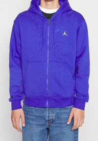 Lila zip-up hoodie i mjukt material. Har framfickor och en logotyp på bröstet. Matchad med blå jeans.