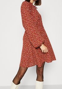 Vestido rojo de flores con mangas largas abullonadas, cintura ajustada y dobladillo con volantes. Presenta pequeñas flores en blanco, amarillo y rosa sobre una tela texturizada.