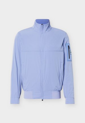 Vaaleansininen vetoketjullinen takki, jossa on korkea kaulus, joustava helmassa, pitkät hihat ja pieni vetoketjullinen tasku vasemmassa ylähiassa.