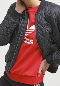 Svart quiltad bomberjacka med ribbade ärmslut, tillsammans med en röd sweatshirt med vit Adidas-logga. Båda plaggen har en avslappnad passform.