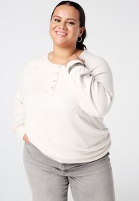 Vrolijke vrouw in een crèmekleurig lange mouwen top en grijze broek, met creolen en armbanden, poserende tegen een effen achtergrond.