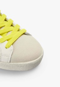 Gros plan sur l'avant d'une basket en daim beige avec une semelle blanche texturée et des lacets jaune vif sur un fond blanc.