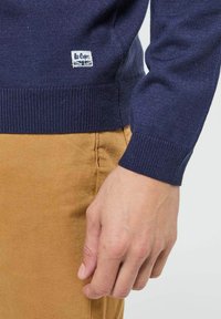 Pull en tricot bleu marine avec poignets et ourlet côtelés, arborant une petite étiquette blanche avec texte et logo. Porté avec un pantalon marron clair.