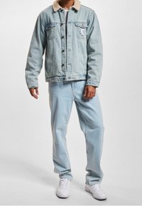 Hombre que lleva una chaqueta de mezclilla azul clara con cuello de borreguito blanco, jeans azul claros a juego y zapatillas blancas, de pie frente a un fondo liso.
