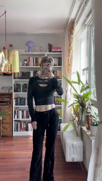 Zwarte langarmige cropped top met een uitsparing, gecombineerd met losse zwarte broeken. De achtergrond bevat boekenplanken en kamerplanten.