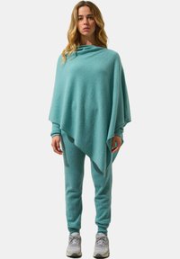 Un poncho verde acqua realizzato in morbido tessuto a maglia, caratterizzato da un design asimmetrico e morbido, abbinato a pantaloni a sigaretta coordinati e sneakers grigie.