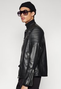 Black leather biker jacket with ribbed shoulder accents and zippered pockets. Worn with a turtleneck and beanie, features a sleek silhouette.