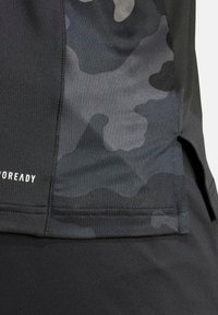 Camiseta atlética negra con un patrón de camuflaje oscuro, textura suave y una abertura lateral. Incluye una marca visible cerca del dobladillo.